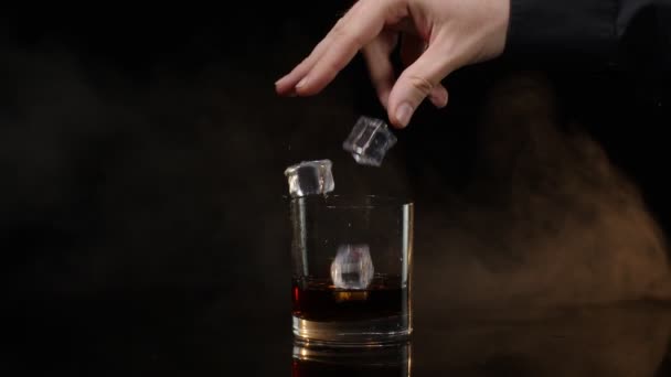 Barman dropping ice cubes into drinking glass with golden whiskey cognac brandy on black background — Stock Video