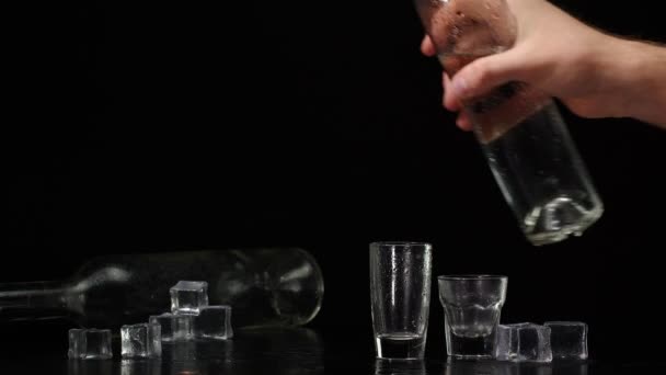 Barman gieten van twee shots van wodka met ijsblokjes uit fles in glazen op zwarte achtergrond — Stockvideo