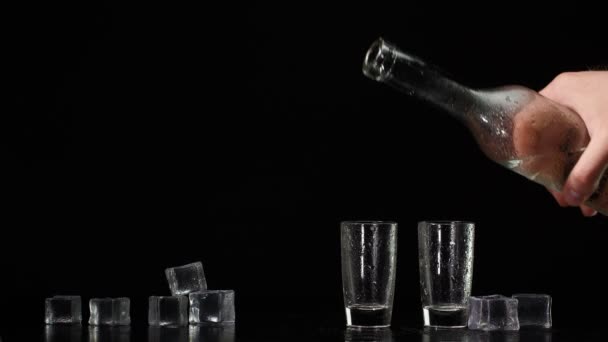 Hand pours vodka, tequila or sake from bottle into shot glasses on black background with ice cubes — Stock Video