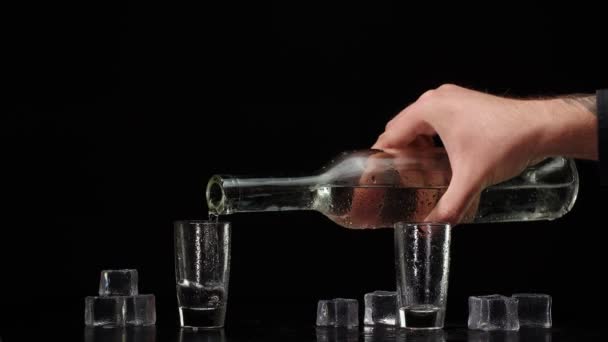 Cantinero vertiendo vodka de botella en dos vasos de chupito con cubitos de hielo sobre fondo negro — Vídeos de Stock