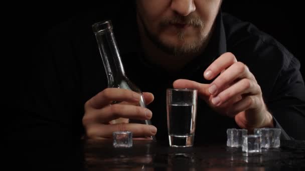 Алкогольний чоловік виливає заморожену горілку з пляшки в склянку з кубиками льоду на чорному тлі — стокове відео