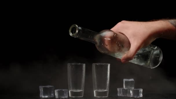 Barman gieten van wodka uit fles in twee shots glazen met ijsblokjes op zwarte achtergrond — Stockvideo