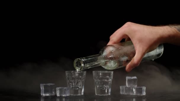 Mão derrama vodka, tequila ou saquê de garrafa em copos de tiro no fundo preto com cubos de gelo — Vídeo de Stock