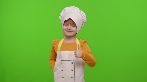 Ребенок девочка одета как повар показывая большие пальцы вверх, улыбаясь, глядя на камеру на хрома ключ — стоковое видео