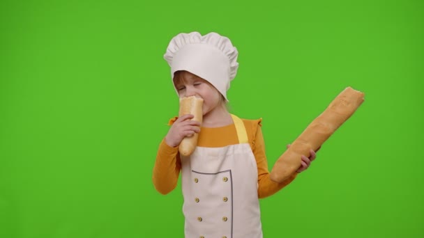 앞치마를 입고 모자를 쓰고 요리사 요리사처럼 갓 만든 맛있는 바게트를 먹고 있는 꼬마 아이 — 비디오