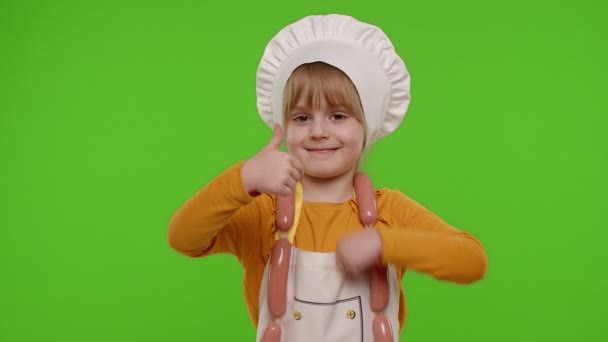 Ребенок девочка одета как повар показывая большие пальцы вверх, улыбаясь, глядя на камеру на хрома ключ — стоковое видео