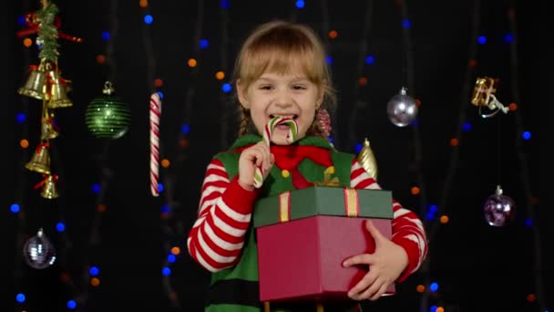 Kid girl in Christmas elf Santa Claus helper costume with gift box licking candy cane lollipop — Stock Video