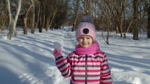 Kind meisje gooien sneeuwbal in de richting van de camera, glimlachend kind lopen, spelen met sneeuw in de winter park — Stockvideo