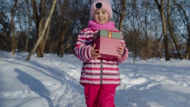 Glimlachend kind in besneeuwde winter bos park op zoek naar camera, met kerstcadeau doos — Stockvideo