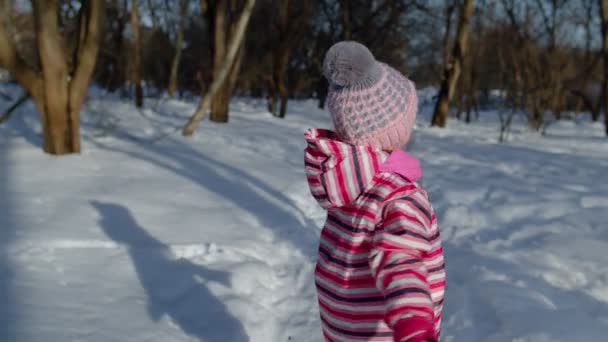 Barnflicka tittar på kameran, ler, dansar, viftar med händerna på snöig väg i vinterparken utomhus — Stockvideo