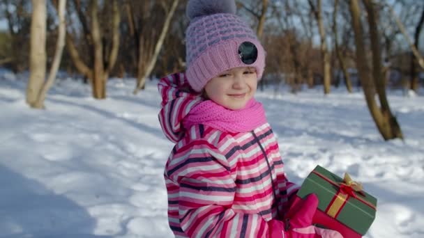 Glimlachend kind in besneeuwde winter bos park op zoek naar camera, met kerstcadeau doos — Stockvideo