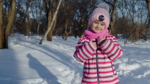 Радісна маленька дівчинка посміхається, показуючи великі пальці вгору жестом на засніженій дорозі в зимовому парку на відкритому повітрі — стокове відео