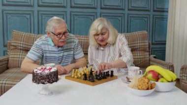 Emekli çift çay içiyor, modern oturma odasında satranç oynuyor.