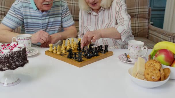 Uśmiechnięta starsza para dziadek babcia spoczywa na kanapie pije kawę, gra w szachy w domu — Wideo stockowe