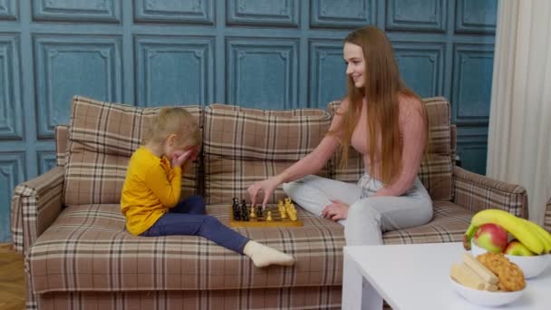 Küçük kız çocuğuna evdeki kanepede tahta tahtada satranç oynamayı öğreten mutlu bir anne kadın. — Stok video