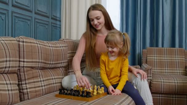 Щаслива молода жінка навчає маленьку дочку дитину грати в шахи на дерев'яній дошці на домашньому дивані — стокове відео