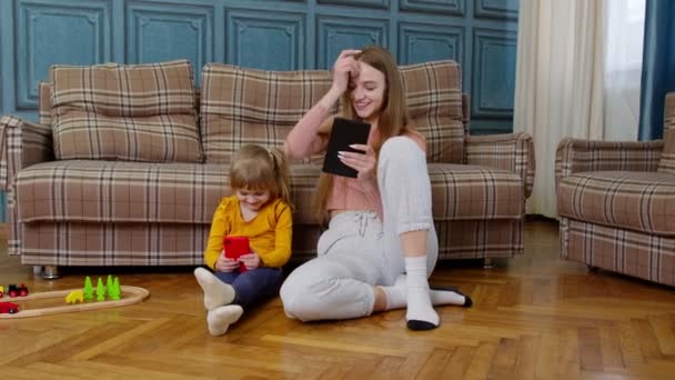 Kvinna barnflicka och barnflicka studerar tillsammans med mobiltelefon och surfplatta, sitter på golvet hemma — Stockvideo