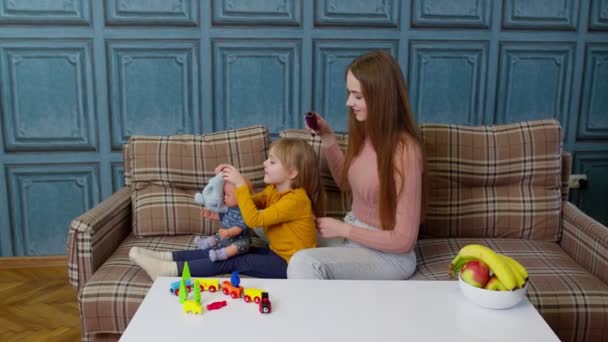 Madre cuidando, cepillando el cabello de la niña hija en la sala de estar, niño jugando con la muñeca de juguete — Vídeo de stock