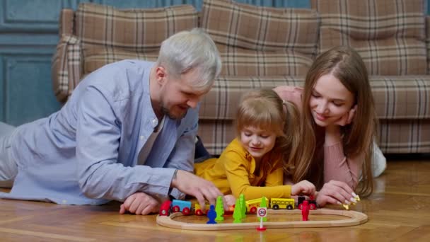 Joven madre y padre jugando con la hija del niño montar tren de juguete en el ferrocarril de madera juego en casa — Vídeos de Stock