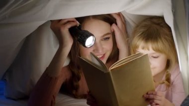 Genç anne, gece odasında yorgan altında kız çocuğuna masal okuyor.
