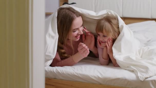 Счастливая семья матери с дочерью лежали на кровати под одеялом, рассказывая секреты друг другу — стоковое видео