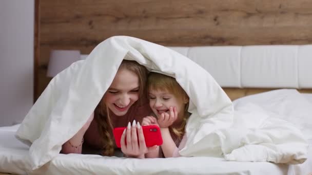 Madre sdraiata sul letto sotto il piumone con figlia bambina che tiene il telefono cellulare guardando film online — Video Stock