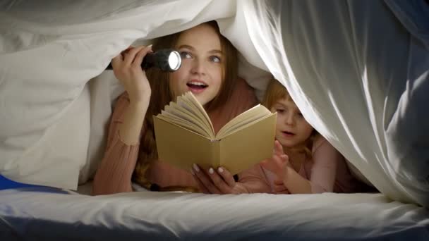 Moeder en dochter liggen thuis in bed onder dekbed deken en lezen verhaaltjes voor het slapen gaan, sprookjes — Stockvideo