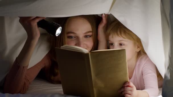 Madre e hija tumbadas en la cama en casa bajo manta nórdica y leyendo cuentos para dormir, cuentos de hadas — Vídeo de stock