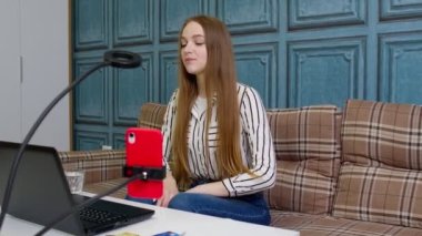 İş kadını blogcusu konuşuyor, cep telefonuyla video blogu kaydediyor, ev ofisinde dizüstü bilgisayar kullanıyor.