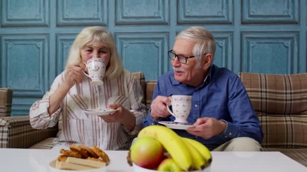 Зріла сімейна пара дідусь, бабуся відпочиває на затишному дивані, насолоджуючись розмовою вдома — стокове відео