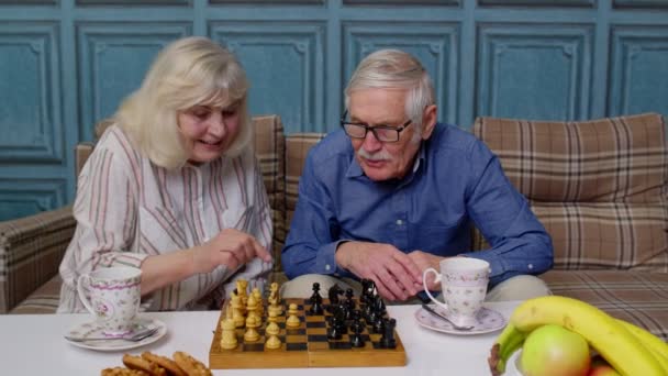 Sonriente pareja de ancianos abuelo abuela descansando en sofá beber café, jugar al ajedrez en casa — Vídeos de Stock