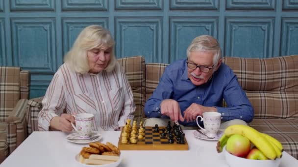 Smiling senior couple grandfather grandmother resting on sofa drinking coffee, playing chess at home — Stock Video