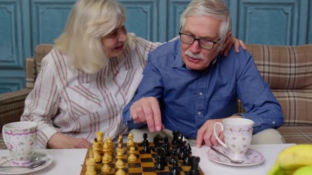 Sonriente pareja de ancianos abuelo abuela descansando en sofá beber café, jugar al ajedrez en casa — Vídeo de stock