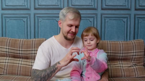 Far og barn datter barn i pyjamas sidde på sofaen smilende, hvilket gør tegn på form hjerte nær brystet – Stock-video