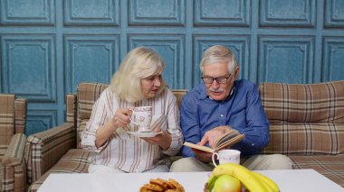 Emekli çift çay içerek, kitap okuyarak modern oturma odasında sohbet ediyor.