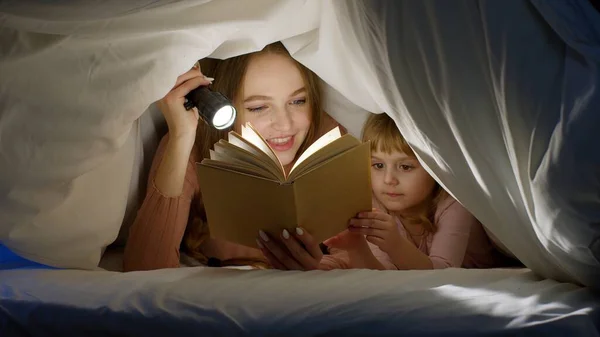 Madre e hija tumbadas en la cama en casa bajo manta nórdica y leyendo cuentos para dormir, cuentos de hadas — Foto de Stock