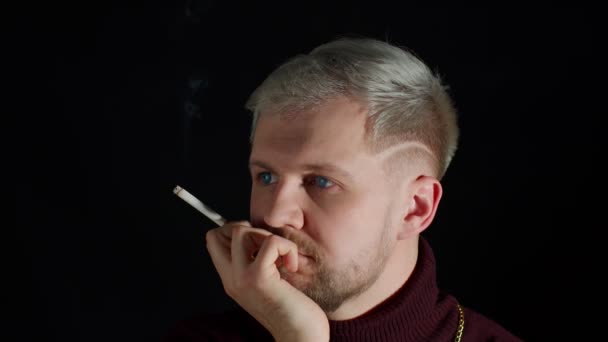 Стильный молодой человек с голубыми глазами в модной одежде смотрит в камеру, курит сигареты — стоковое видео