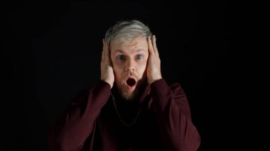 Şaşırmış stil sahibi bir adam heyecan duyuyor, kameraya bakıyor ve yüzüne yakın el ele tutuşuyor.