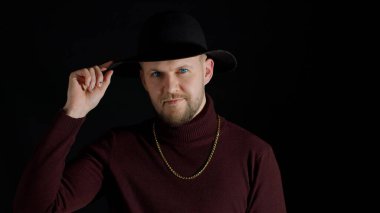 Baştan çıkarıcı gülümseyen, modaya uygun giyinen ve siyah arka planda şapka takan genç bir adamın portresi.