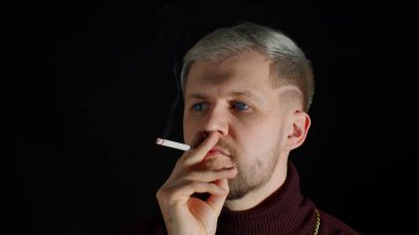 Moda giysileri içindeki mavi gözlü şık bağımlı genç adam kameraya bakıyor, sigara içiyor.