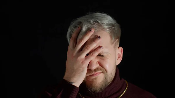 Disappointed man doing face palm gesture on black background. Stressed guy covering face with hand