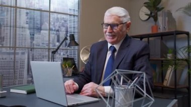 Neşeli kıdemli şirket yöneticisi dizüstü bilgisayarda çalışıyor, gülüyor, zaferi kutluyor.