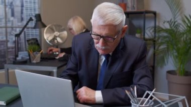 Ofiste çalışan yaşlı bir patron çenesini ovuyor, çözümü düşünüyor, şüpheli sorular soruyor.