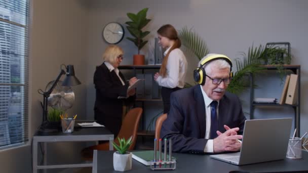Веселый старший бизнесмен, работающий на портативном ноутбуке, сидит за столом, слушает музыку и танцует — стоковое видео
