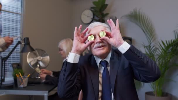 Starší pán tančí, fláká se, dělá hloupé obličeje s bitcoinovýma očima v kanceláři — Stock video