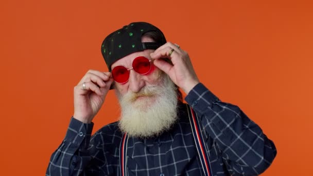 Boldog játékos idős szakállas férfi napszemüvegben és sapkás pislogó szemmel, mosolyogva néz a kamerába — Stock videók