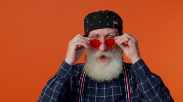 Vieil homme barbu âgé séduisant portant des lunettes de soleil, sourire charmant, pointant vers la caméra — Video