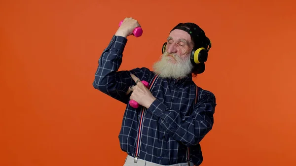 Пожилой бородатый мужчина слушает музыку через наушники, тренируется, поднимает розовые гантели, здравоохранение — стоковое фото