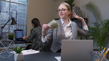 Gülümseyen genç iş kadını ofis masasında yığınla parayla el sallıyor.