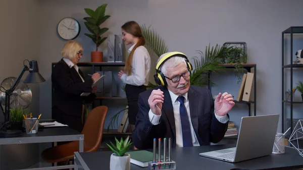 Χαρούμενος ανώτερος επιχειρηματίας που εργάζεται σε φορητό φορητό φορητό φορητό υπολογιστή κάθεται στο γραφείο, ακούγοντας μουσική και χορό — Φωτογραφία Αρχείου